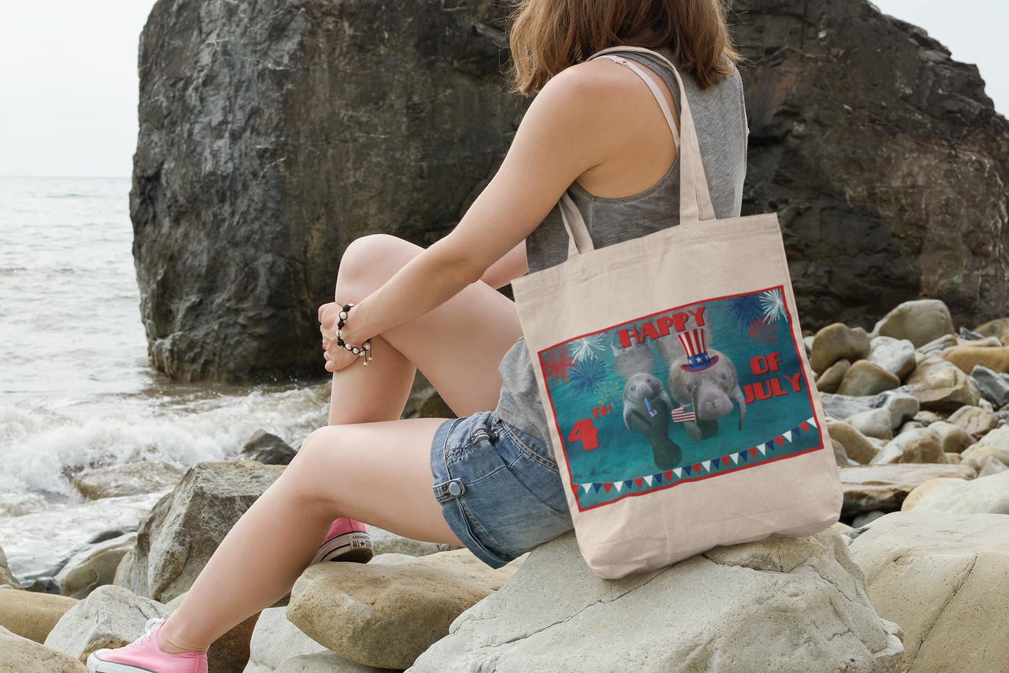 Happy 4th of July Manatee Tote Bag, model sitting on rocks by ocean
