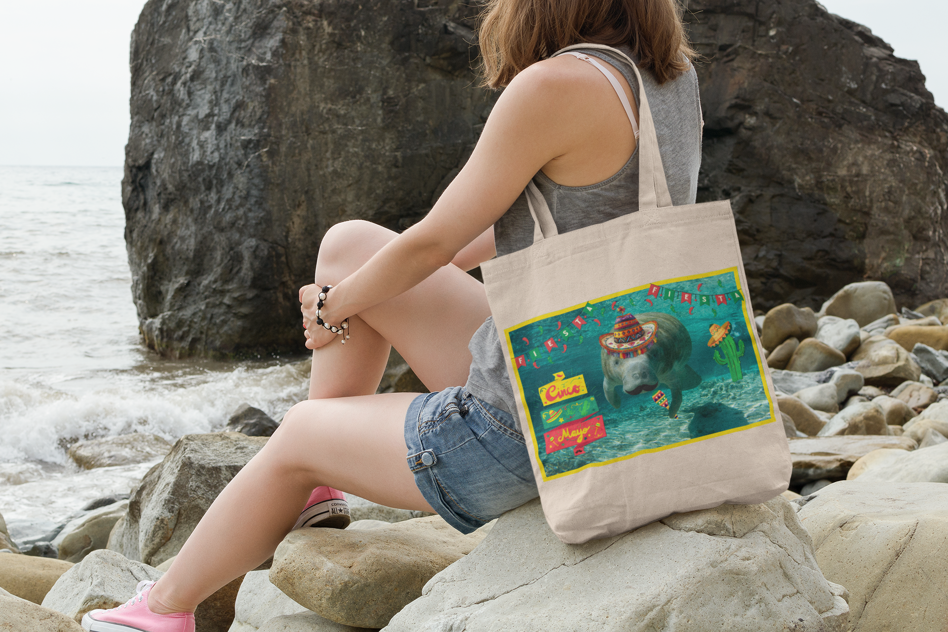 Happy Cindo de Mayo Manatee Tote Bag, model sitting on rocks by ocean