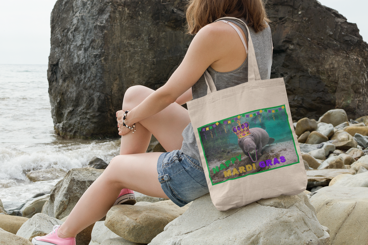 Happy Mardi Gras Manatee Tote Bag, model sitting on rocks by ocean