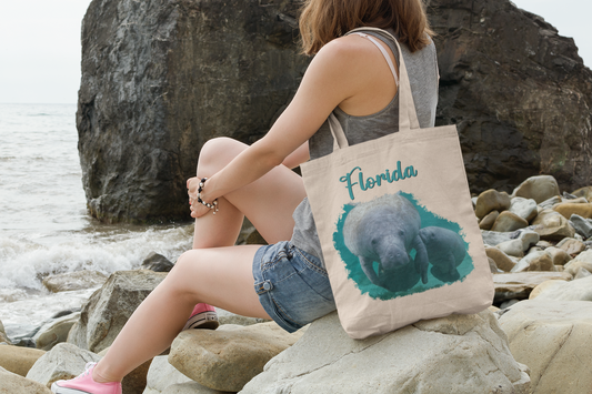 Manatee and Calf Feeding Tote Bag, model with tote sitting on rocks by ocean