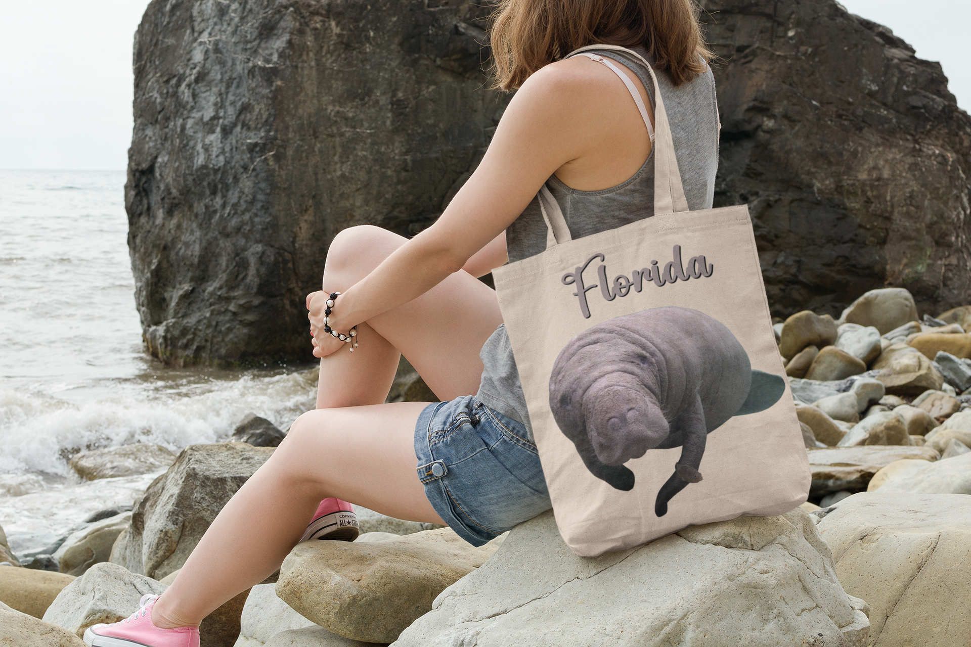 Manatee and Little Fish Tote Bag, model with tote sitting on rocks by ocean