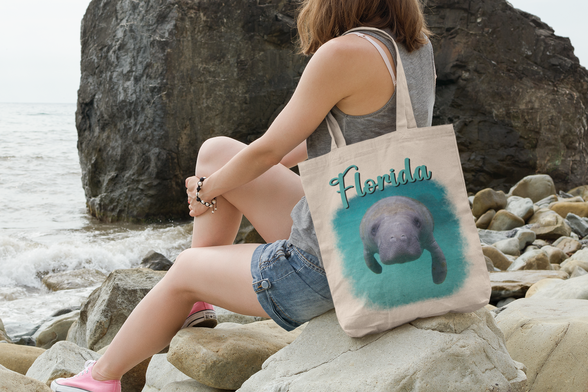 Curious Manatee Tote Bag, model with tote sitting on rocks by ocean