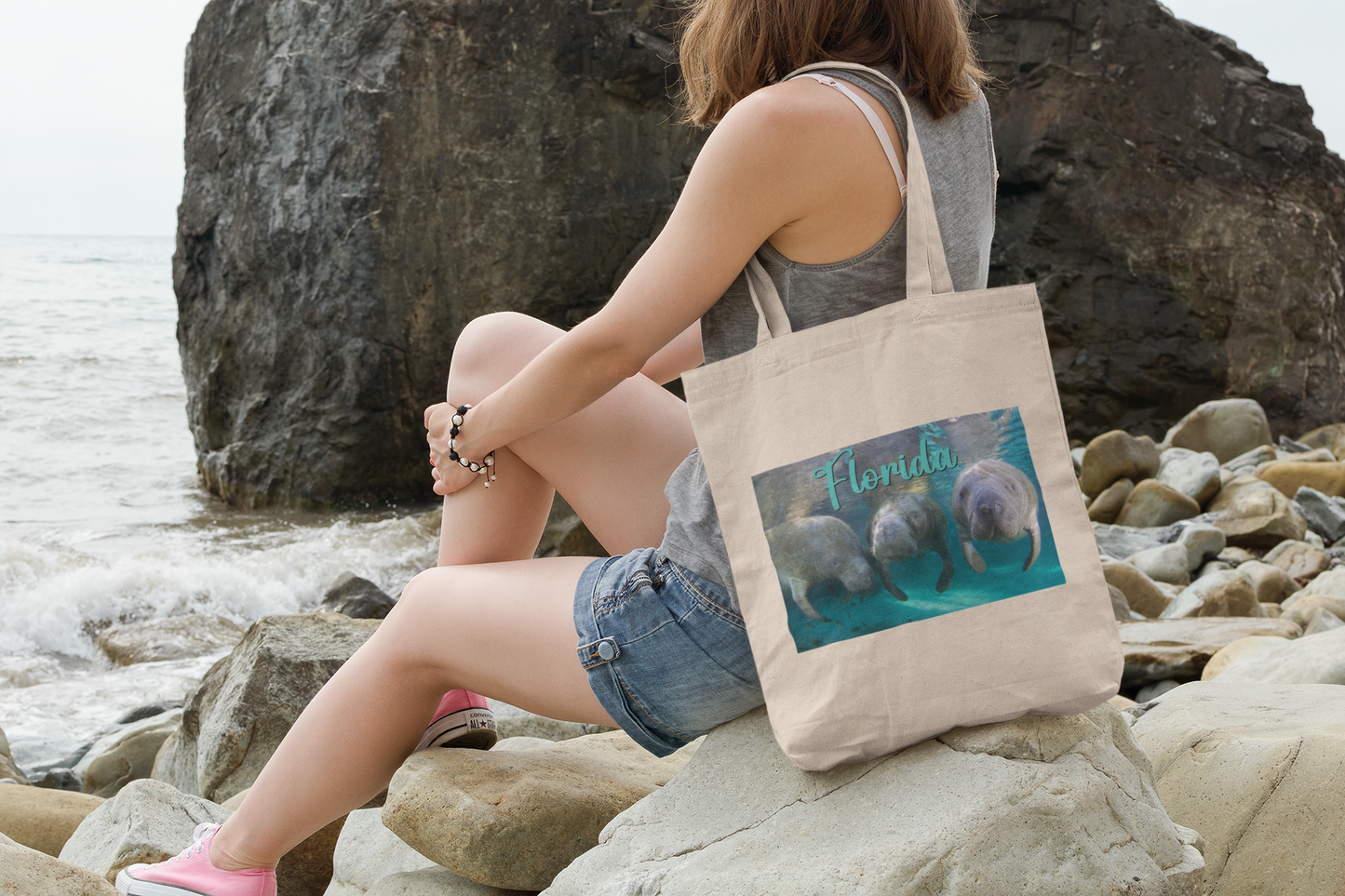 3 Manatees Tote Bag, model with tote sitting on rocks by ocean