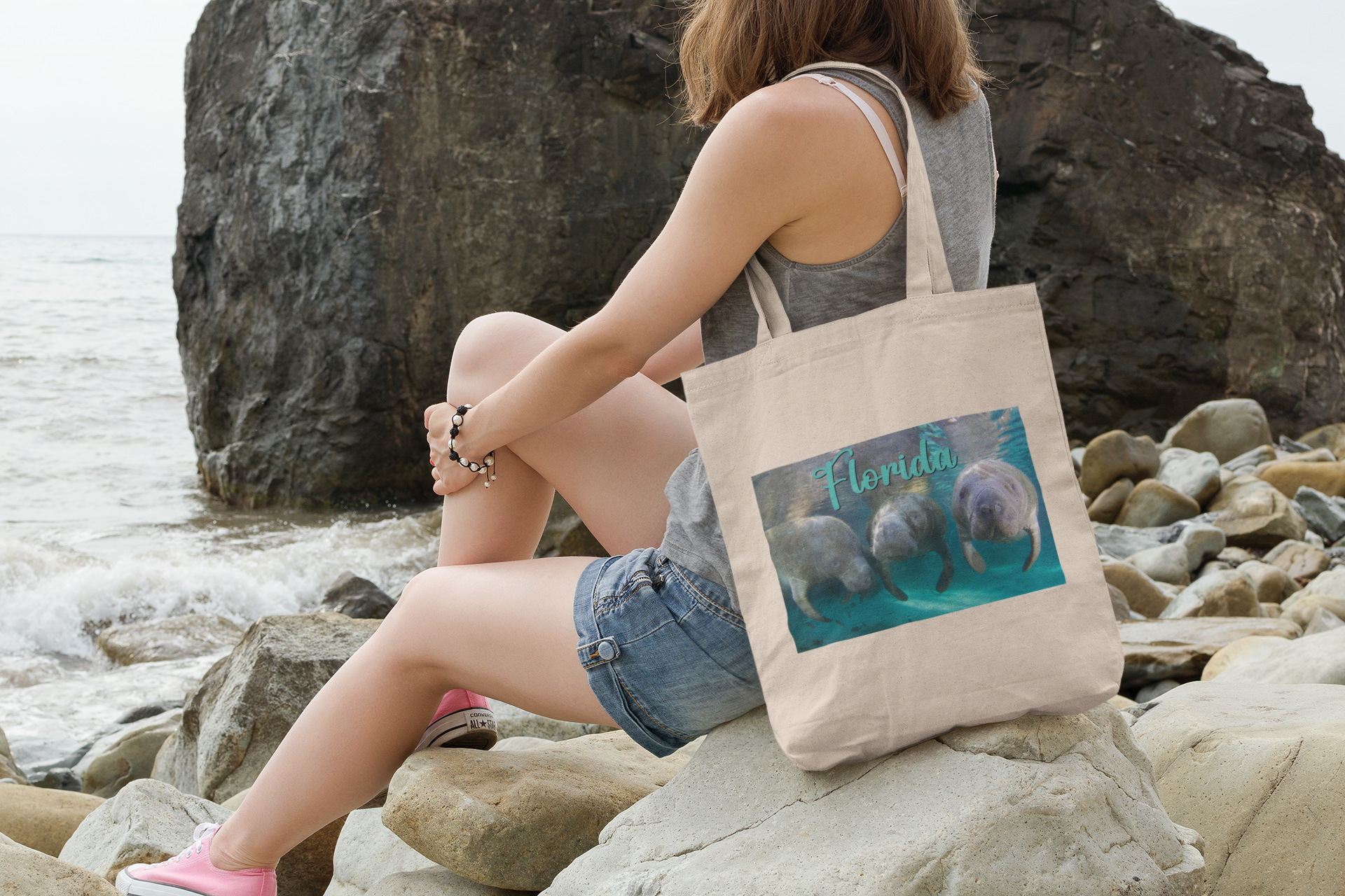 3 Manatees Tote Bag, model with tote sitting on rocks by ocean