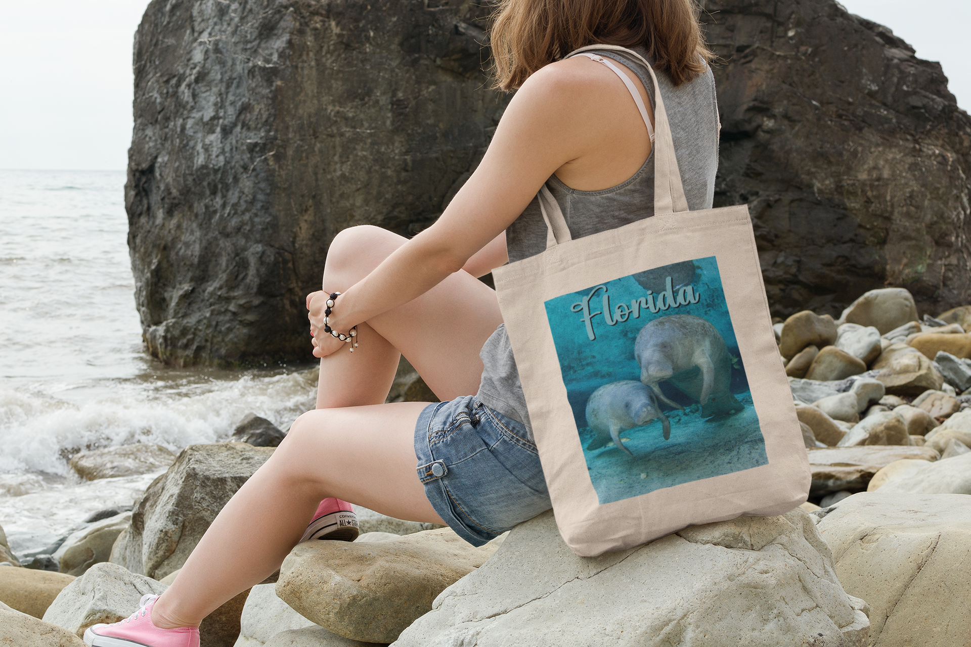 2 Graceful Manatees Tote Bag, model with tote sitting on rocks by ocean
