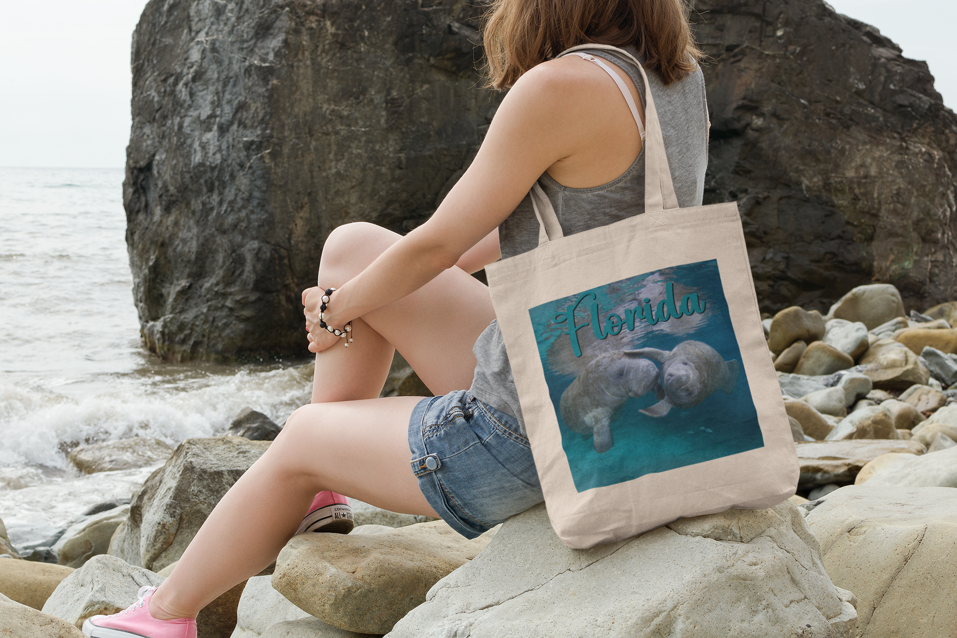 Playful Manatee Tote Bag, model with tote sitting on rocks by ocean